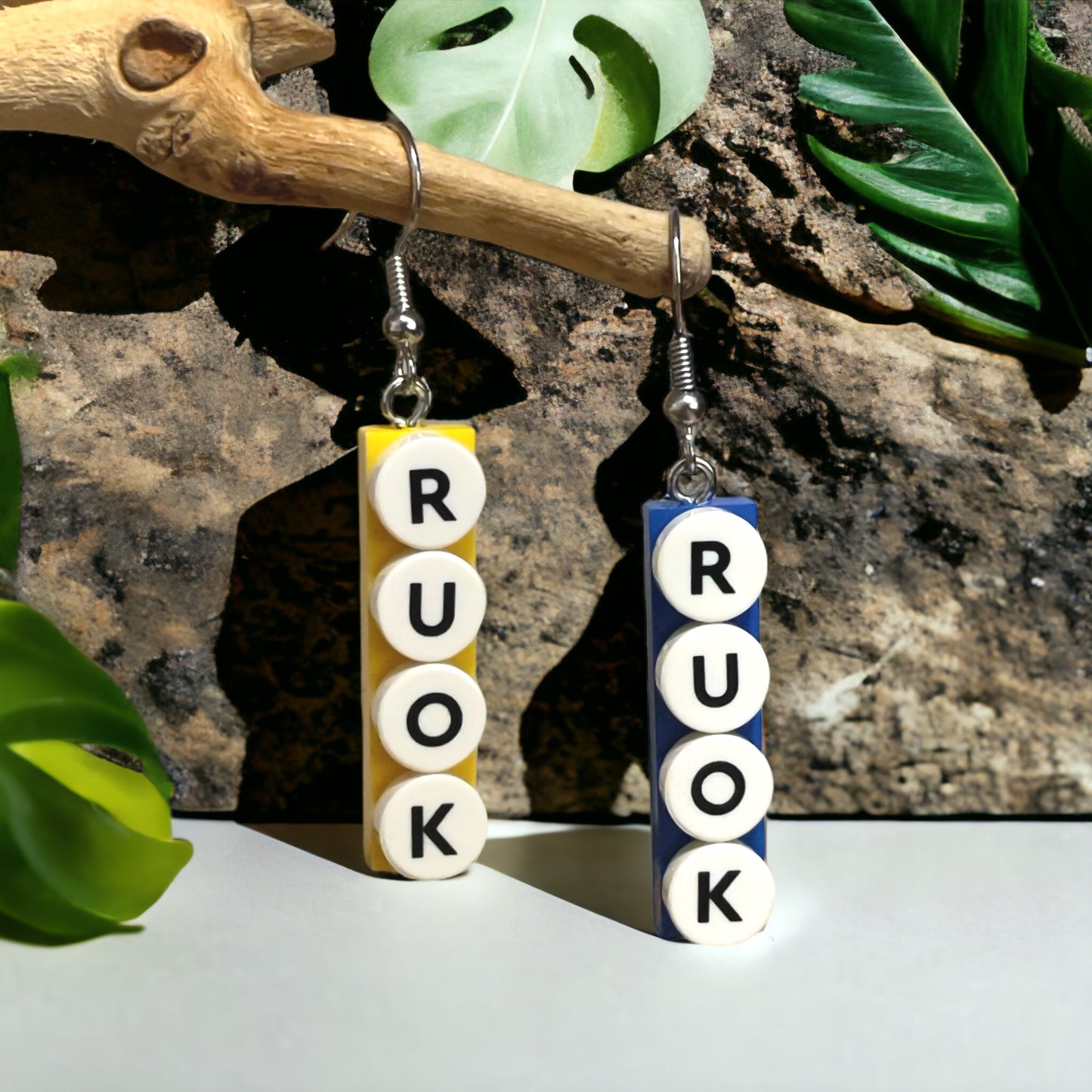 A blue and a yellow Lego plate with black letters printed ‘R U OK’ on round white pieces placed vertically down the plates as hook earrings. They are hanging on a branch above a smooth white surface with a rock face and three green leaves in the background.