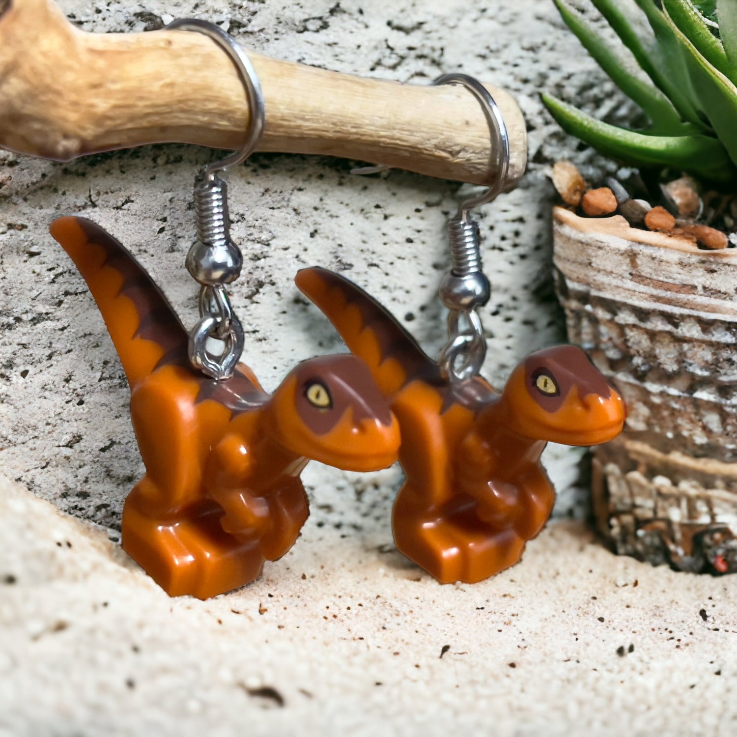 Two light brown Lego t-rex style dinosaurs with a dark brown strip from their heads to tails as hook earrings. They are hanging off a branch in front of a textured grey surface with a plant and pebbles in a brown pot on the right hand side in the background.