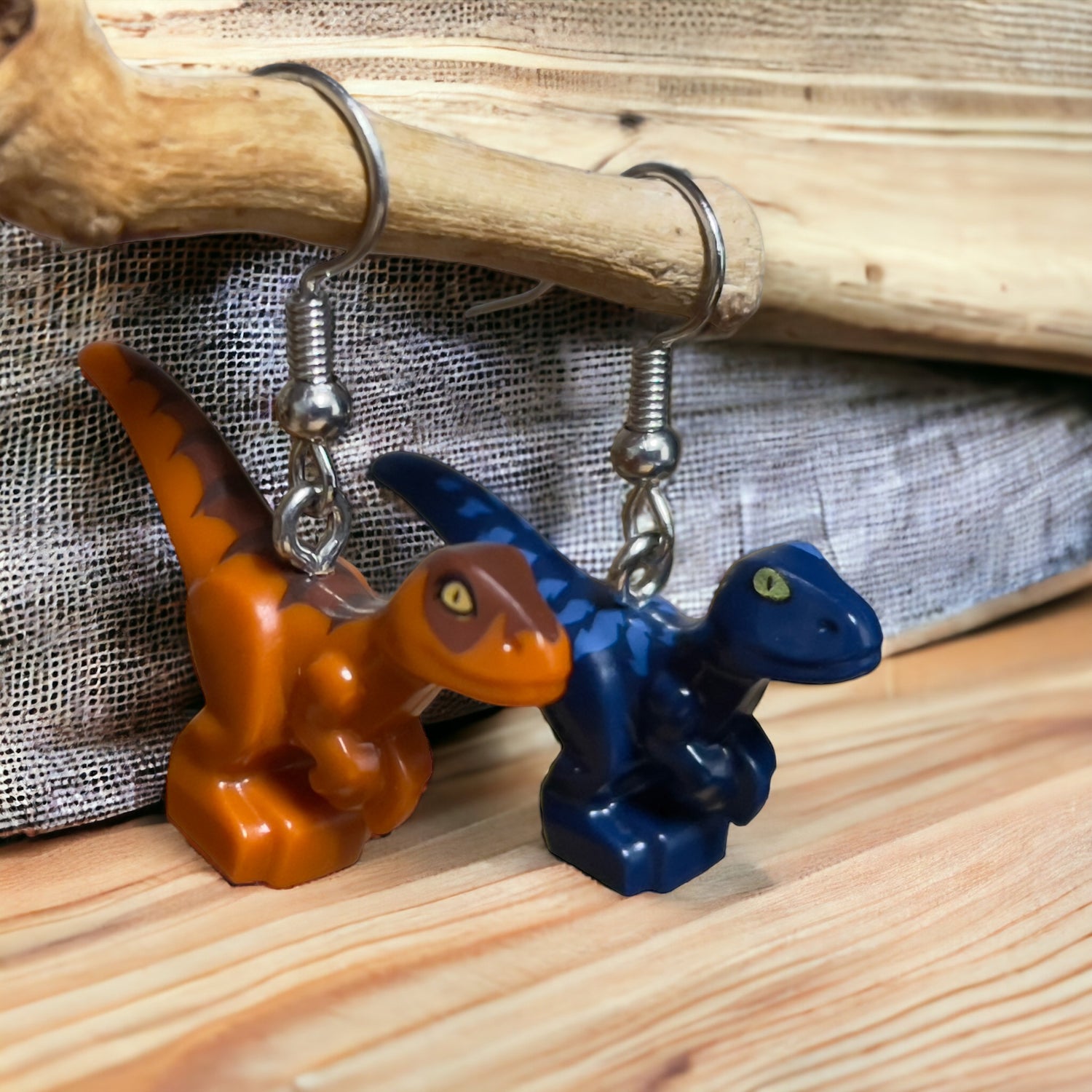 Two mismatched Lego t-rex style dinosaurs as hook earrings. One dinosaur is light brown with a dark brown strip from its head to tail and the other dinosaur is dark blue with light blue stripes down its back. They are hooked on a branch while standing on a light oak wood grain surface with textured grey fabric behind
